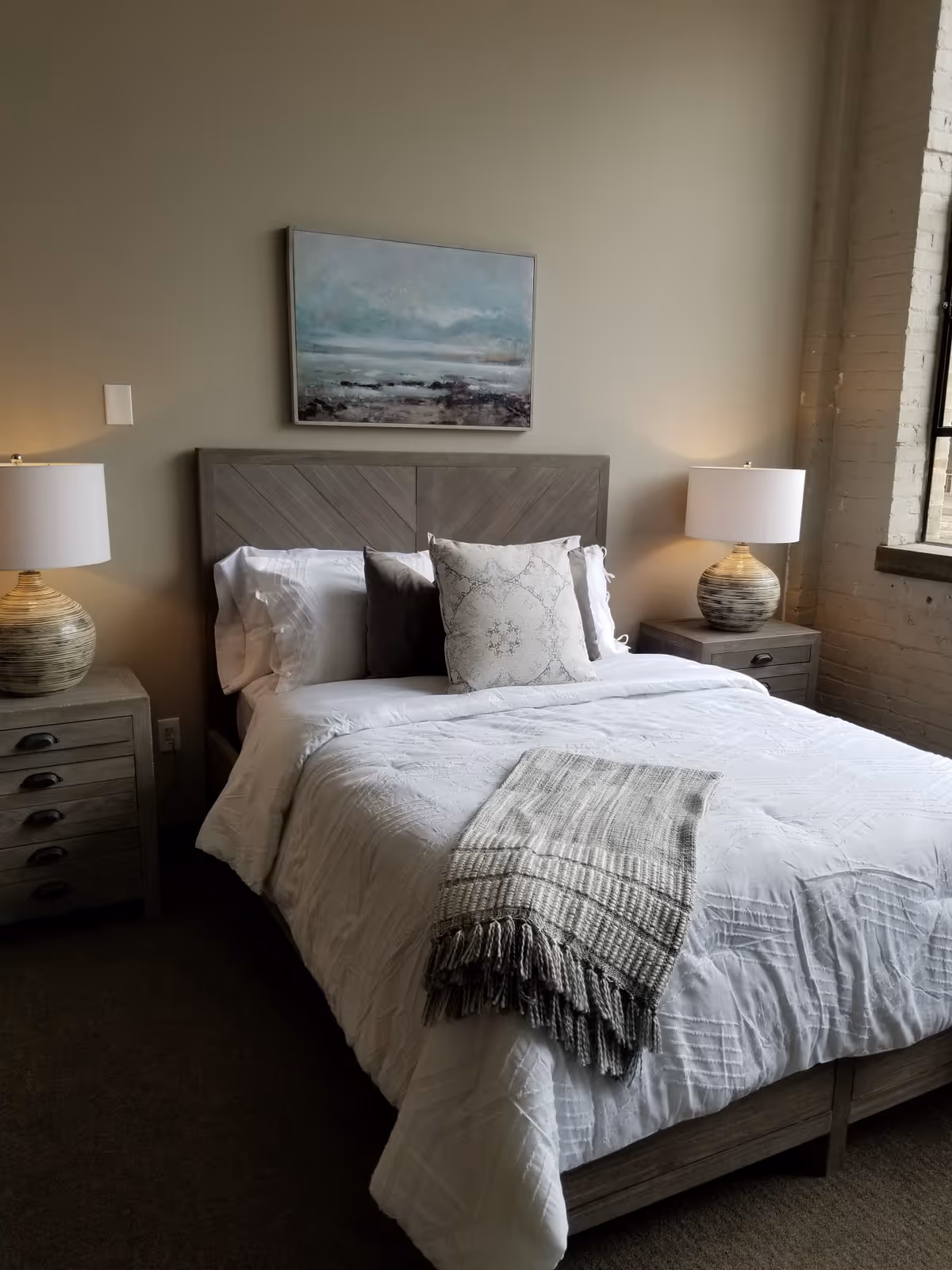 A neatly made bed with white bedding and decorative pillows in a bedroom. There is a gray wooden headboard, two matching nightstands with lamps on each side, and a framed landscape painting hanging above the bed. A window with a brick wall is visible on the right side.