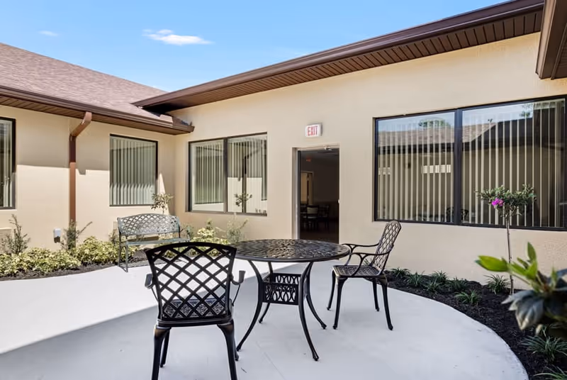 Outdoor patio area at Gold Choice Palm Coast Assisted Living & Memory Care with a round metal table and three matching chairs on a concrete surface. The patio is surrounded by beige building walls with windows and a door marked with an exit sign. There are small plants and shrubs along the building walls.