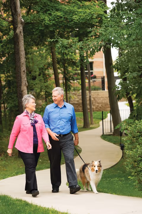 An elderly couple walking a dog on a paved path surrounded by green trees and grass in an outdoor setting near a building.