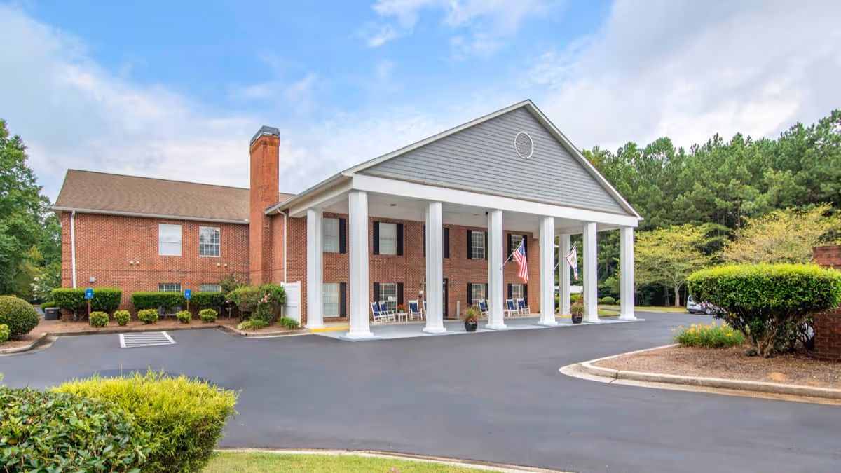 Front entrance of a two-story brick senior living building with white columns, flags, and a paved driveway.