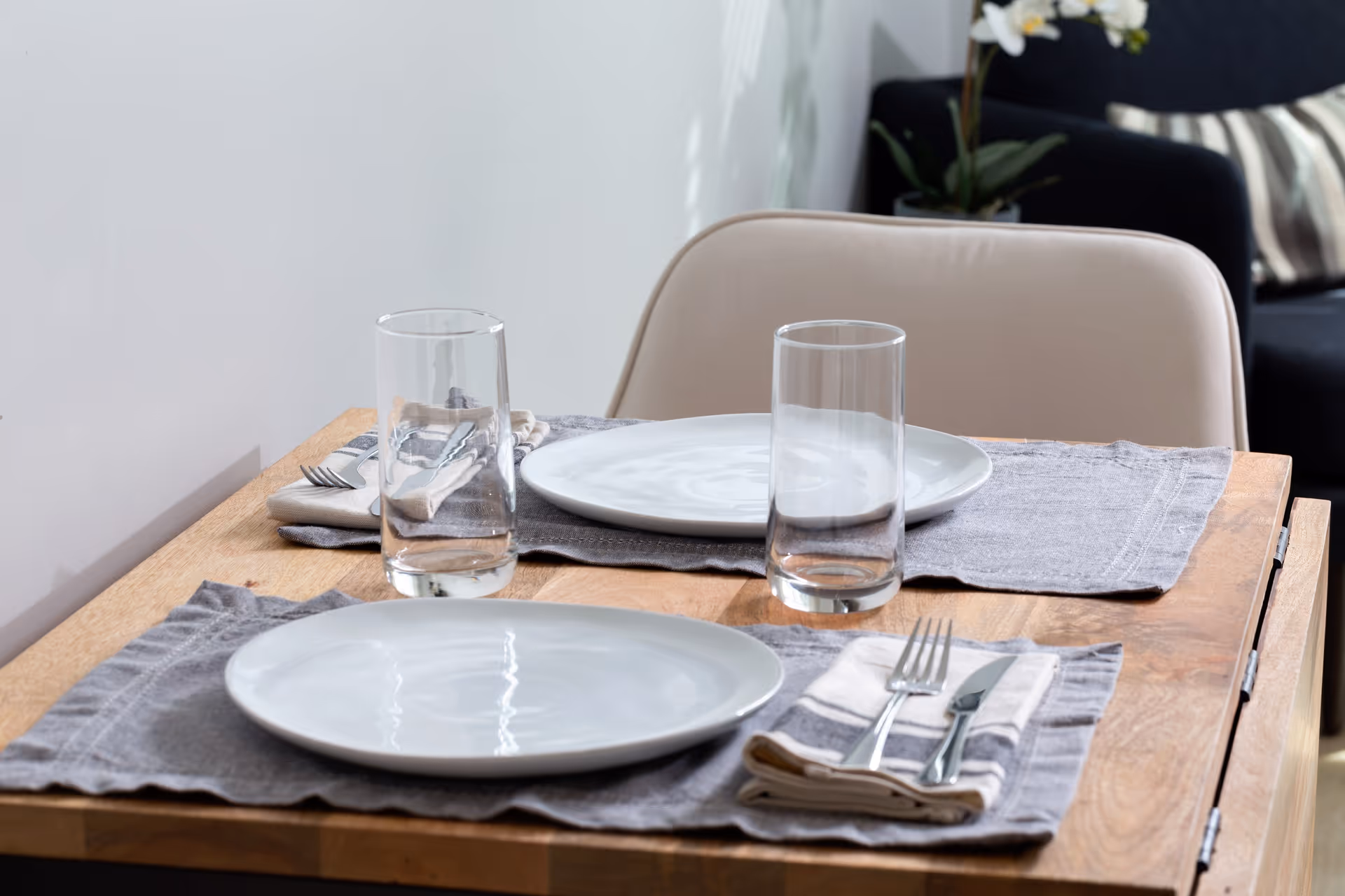 A small wooden dining table set for two with white plates, clear drinking glasses, and silverware wrapped in cloth napkins on gray placemats. A beige chair is positioned at the table, and a dark armchair with a striped pillow and a potted orchid are visible in the background.