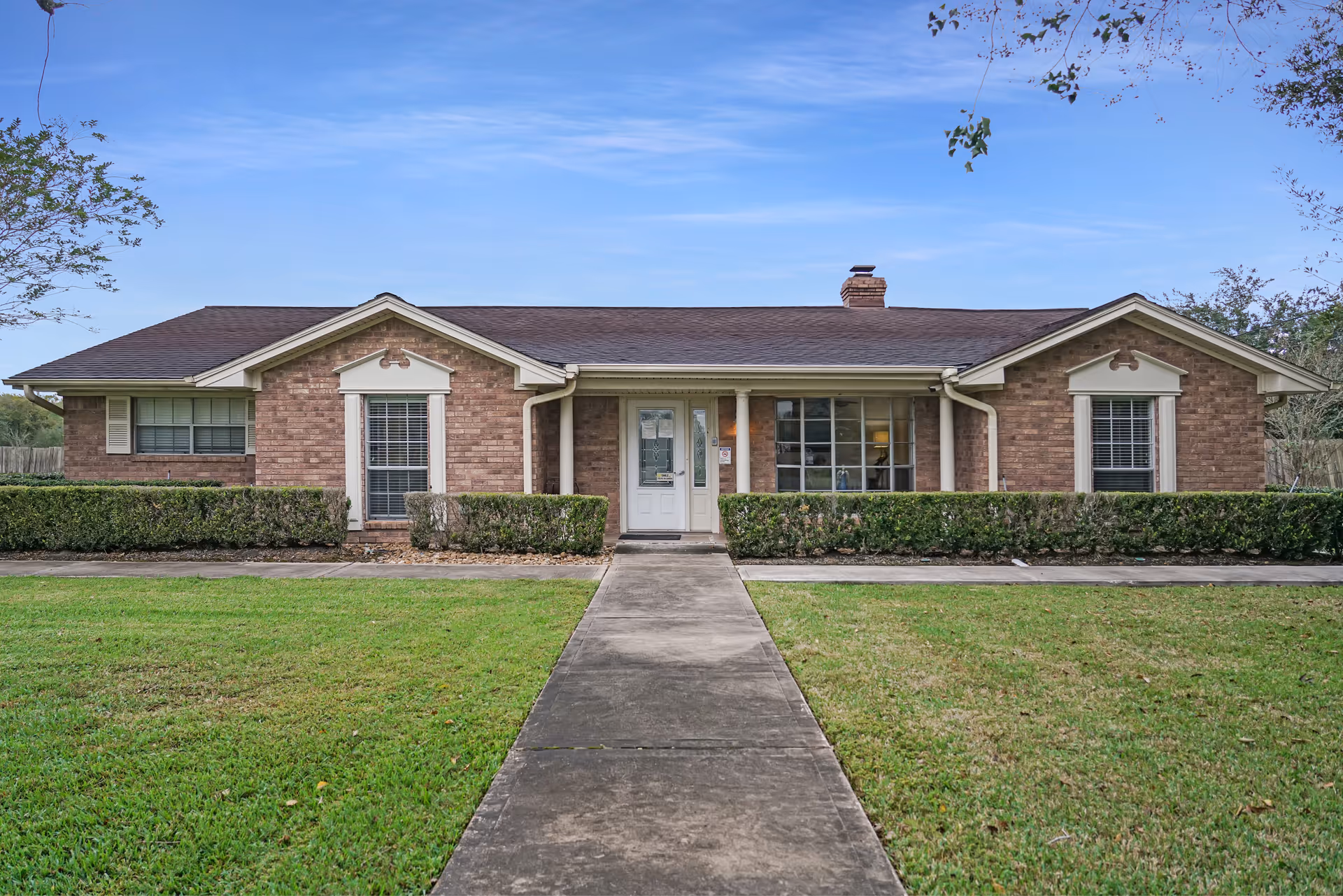 Single-story brick building with a central walkway, manicured lawn and hedges leading to a white front door and windows.