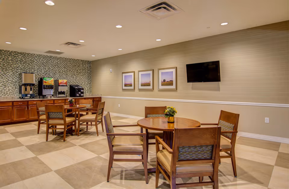 A dining area with round wooden tables and chairs, a countertop with beverage dispensers, a wall-mounted TV, and three framed pictures on the wall. The floor has a checkered tile pattern and the walls are painted beige with a decorative tile backsplash behind the countertop.
