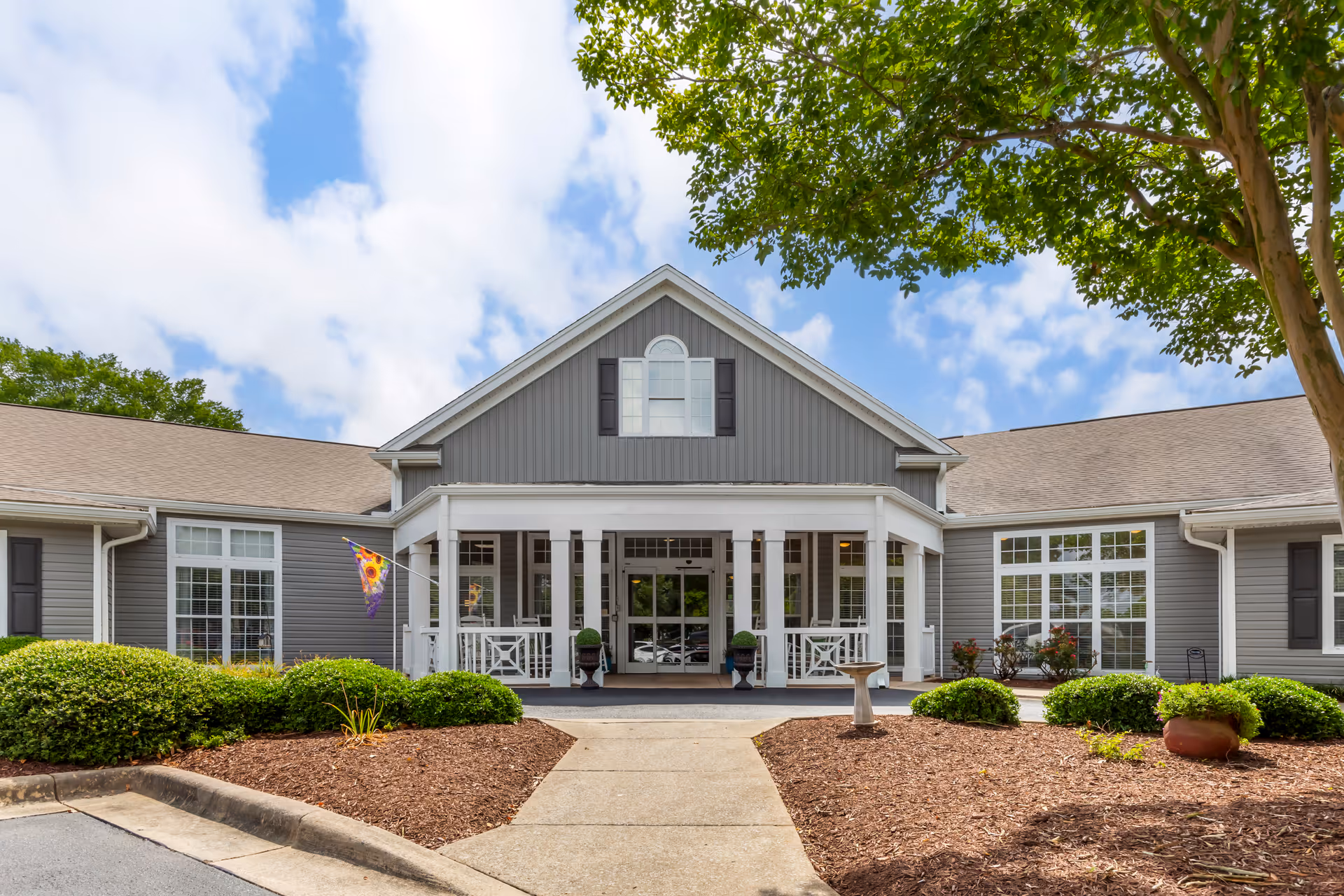 Front exterior view of a single-story senior living facility building with gray siding, white trim, large windows, a covered entrance with white railings, and landscaping with bushes and a tree under a partly cloudy sky.