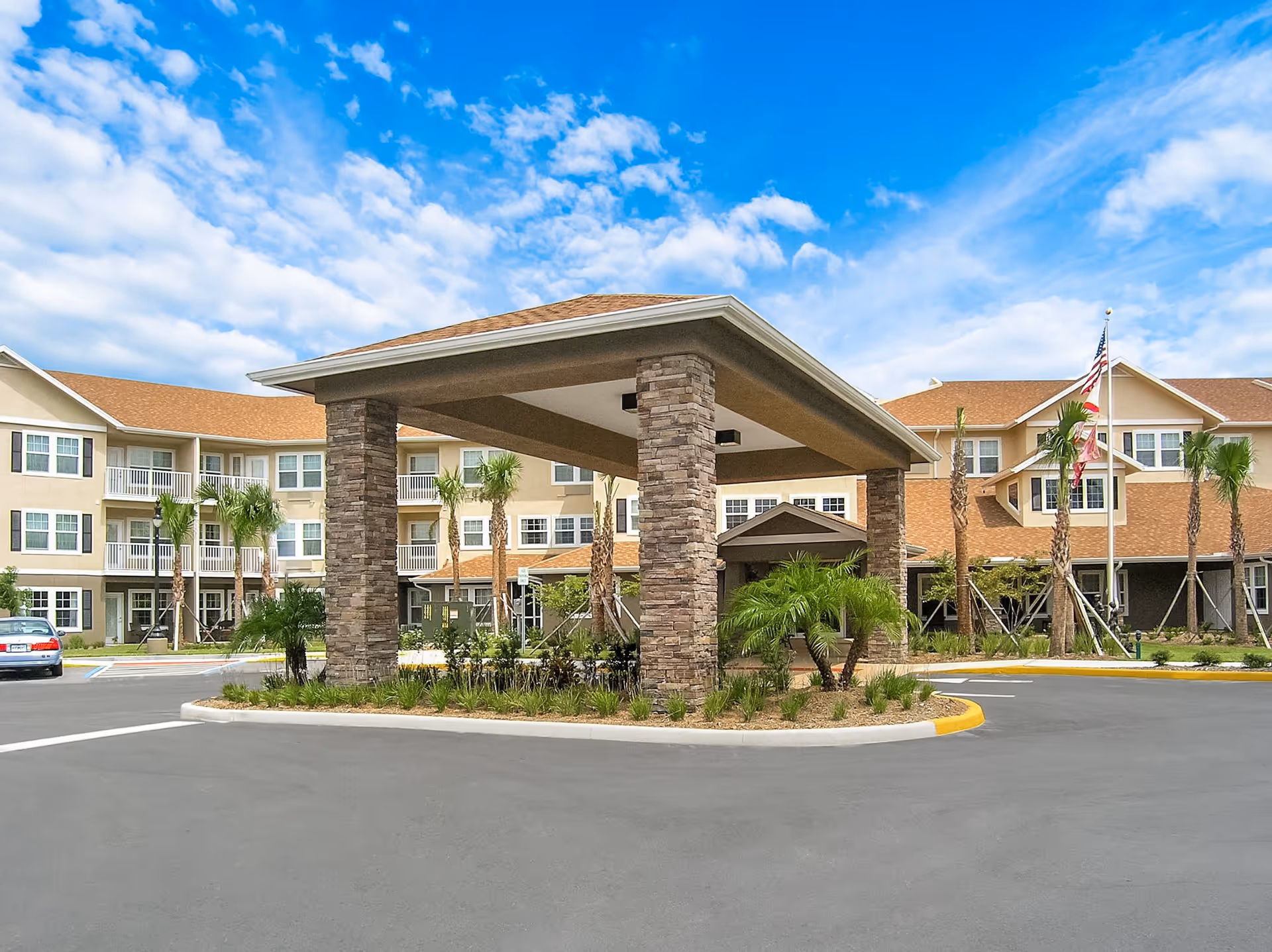 Exterior view of The Windsor of Palm Coast facility showing a covered entrance with stone pillars, surrounded by a well-maintained driveway, palm trees, and a three-story building with beige walls and multiple windows under a blue sky with scattered clouds.
