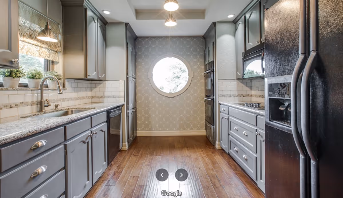 A modern kitchen with gray cabinets, granite countertops, and wooden flooring. The kitchen features a double sink under a window with potted plants, a black dishwasher, a black refrigerator with a water dispenser, a built-in oven, a microwave, and a stovetop. A round window at the far end lets in natural light, and there are pendant lights hanging from the ceiling.