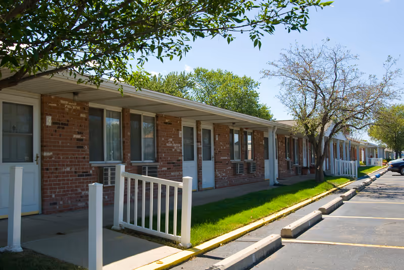 Exterior view of a single-story brick building with multiple doors and windows, each unit having a small porch area with white railings. There is a parking lot with marked spaces in front of the building and trees providing some shade.