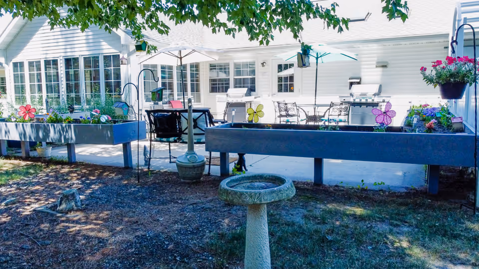 Patio area with raised planter boxes, chairs, umbrellas and a birdbath in front of a white senior living building.