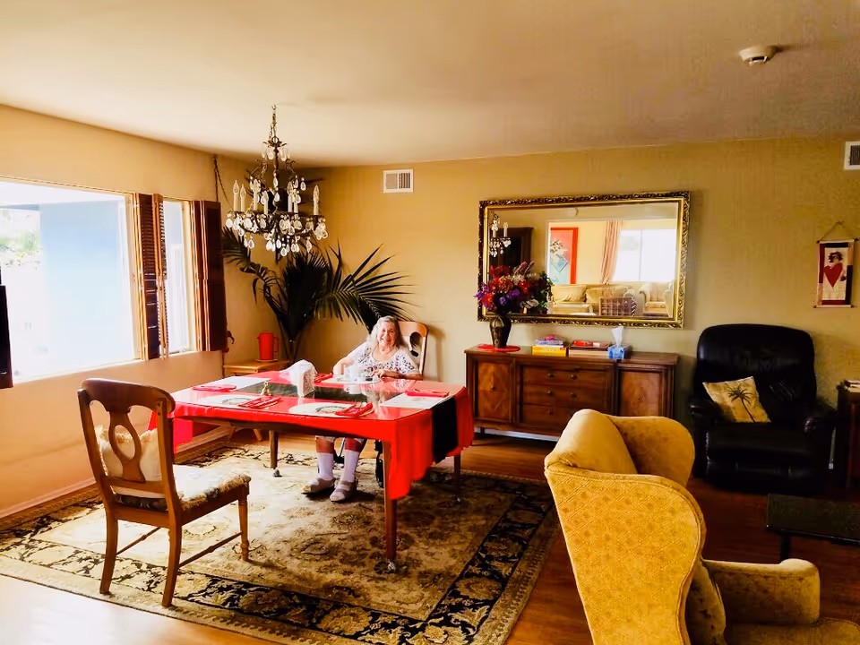 A cozy living room and dining area with a large window letting in natural light. A woman is sitting at a dining table covered with a red tablecloth, set with placemats and napkins. The room features a chandelier, a large decorative mirror above a wooden sideboard with books and a flower vase, a black armchair with a palm tree pillow, and a yellow upholstered chair. The floor has a patterned rug and wooden flooring.