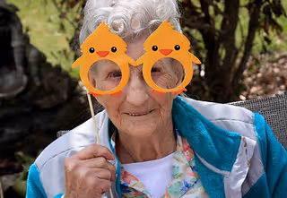 An elderly woman outdoors holding up a pair of orange novelty glasses shaped like birds in front of her eyes, smiling gently. She is wearing a light blue jacket over a floral shirt, with greenery and a stone feature in the background.