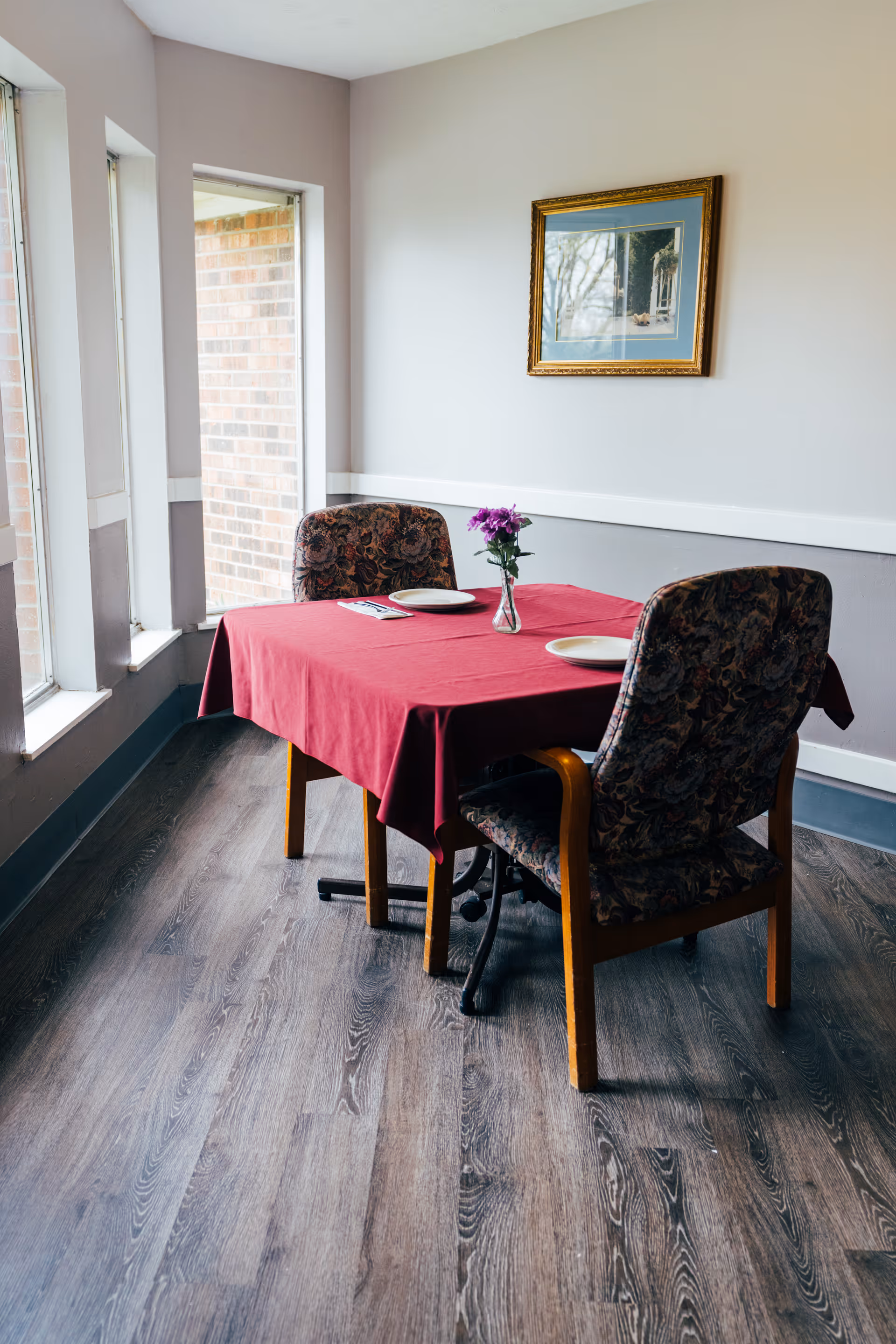 Small dining nook with a red tablecloth on a two-person table, upholstered chairs, a vase of flowers, and a framed picture on the wall.