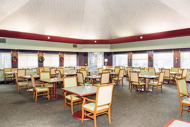 A spacious dining room with multiple square tables and wooden chairs with light green cushions. The room has large windows with white blinds and holiday wreath decorations. The ceiling is high and dome-shaped with a maroon border near the top of the walls.
