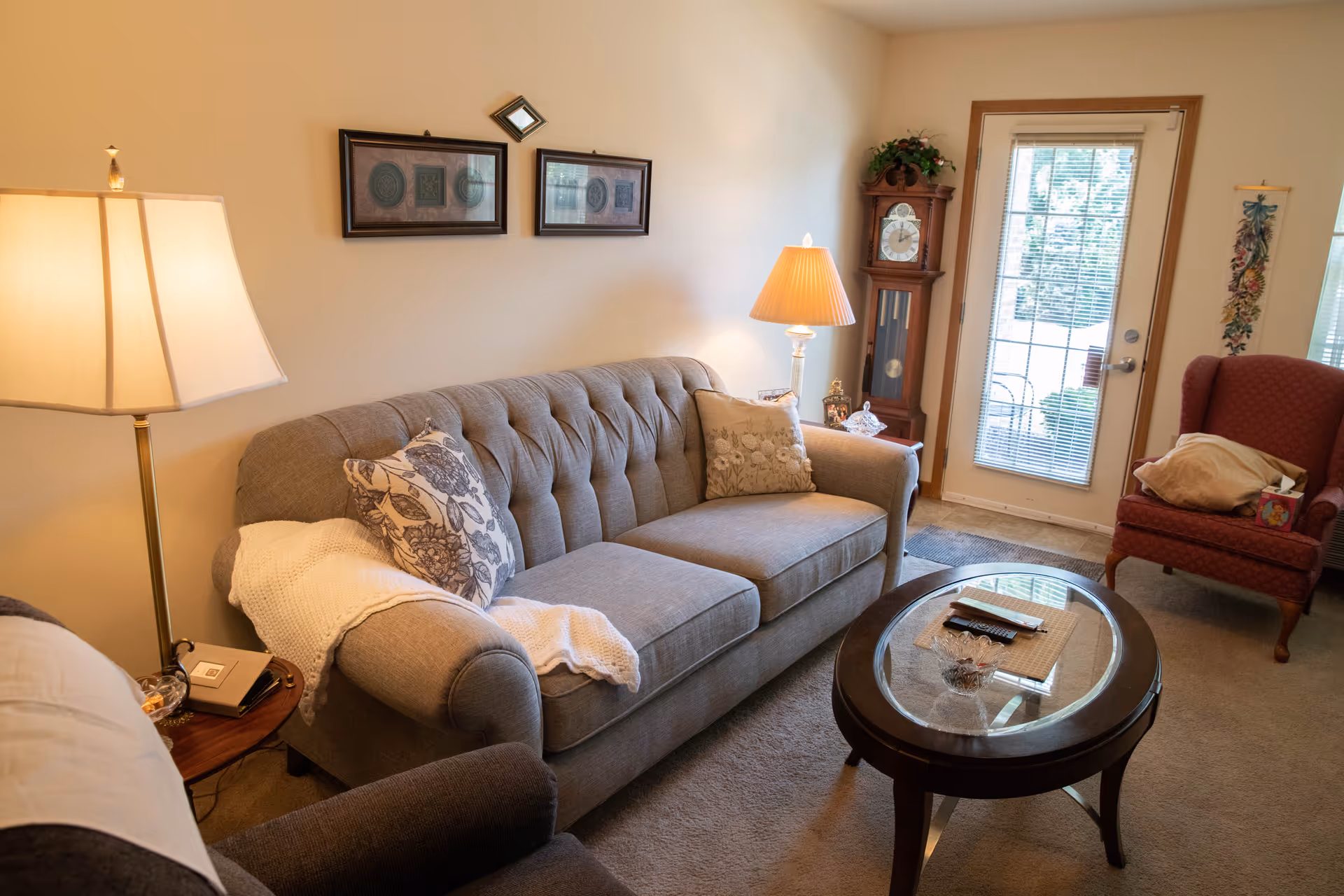 Cozy living room with a gray tufted sofa adorned with patterned pillows and a white throw blanket. A wooden side table with a lamp and a phone sits next to the sofa. Across from the sofa is a round glass-top coffee table with a remote control and decorative items. In the background, there is a tall grandfather clock, a red armchair with pillows, and a door with glass panes and blinds leading outside. The walls are decorated with framed artwork and a small mirror.