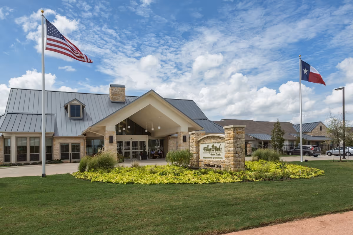 Front exterior view of the Village on the Park Onion Creek facility with a stone entrance sign, two flagpoles displaying the American and Texas flags, a well-maintained lawn, and a partly cloudy sky.