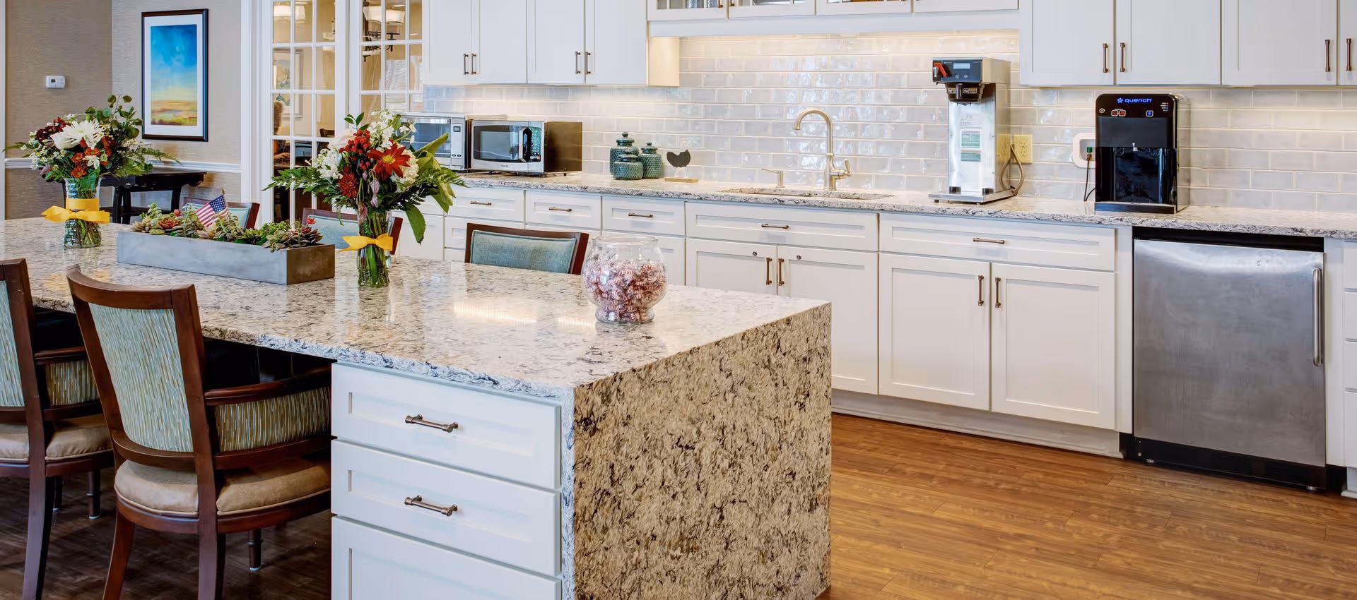 Bright communal kitchen with a large granite island, seating, white cabinets, stainless dishwasher and a coffee station.