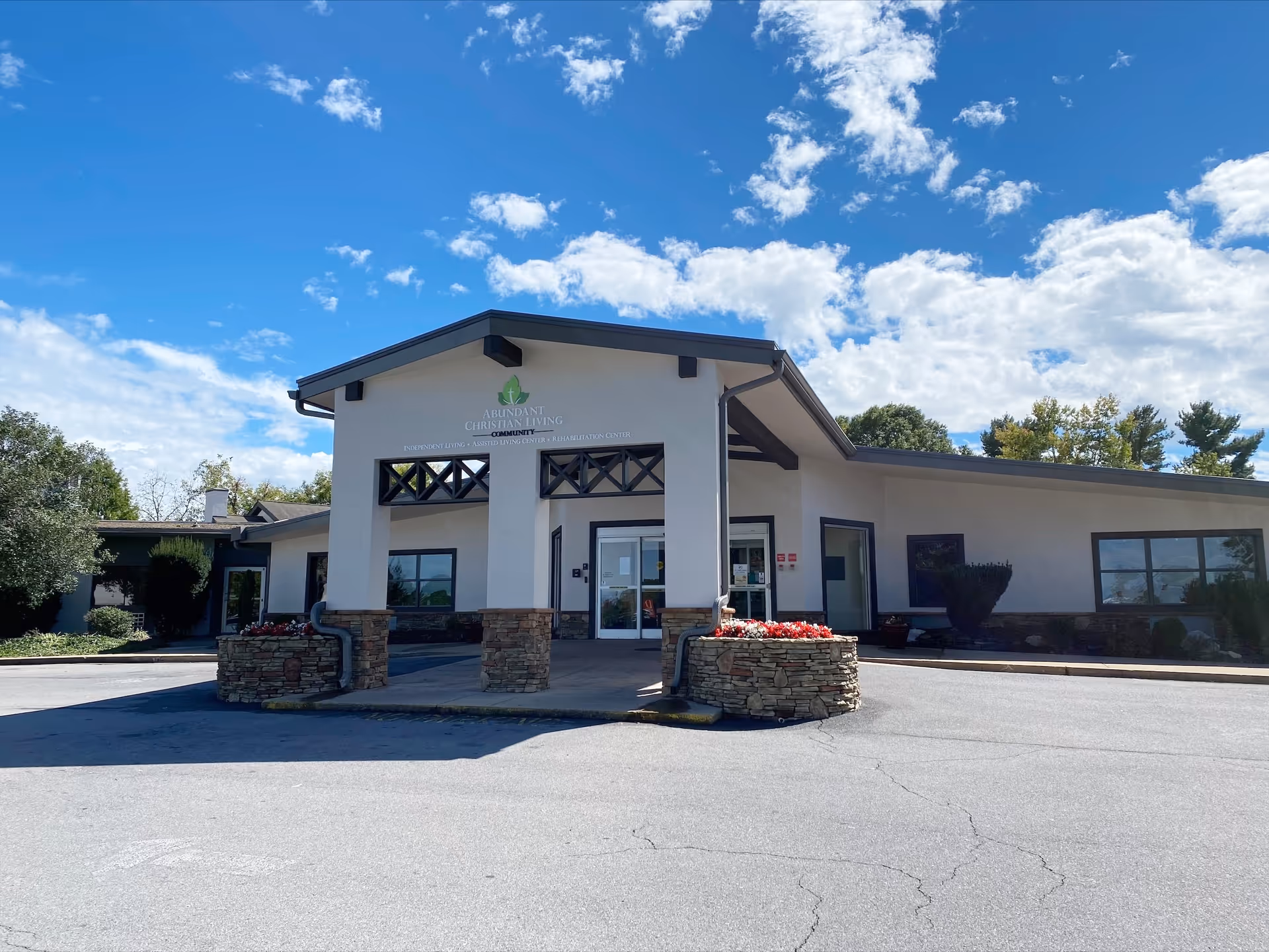 Front exterior view of Abundant Christian Living Community building with a covered entrance, stone planters with flowers, and a clear blue sky with scattered clouds.