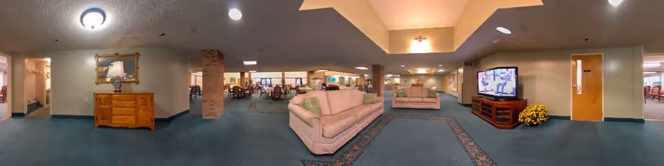 A spacious senior living facility common area with blue carpet, beige walls, and ceiling lights. The room features two beige sofas with green pillows arranged around a patterned rug. A wooden TV stand holds a flat-screen television, and a yellow flower arrangement is placed beside it. In the background, there are dining tables and chairs visible through open doorways, and a wooden dresser with a lamp and framed mirror is on the left side.
