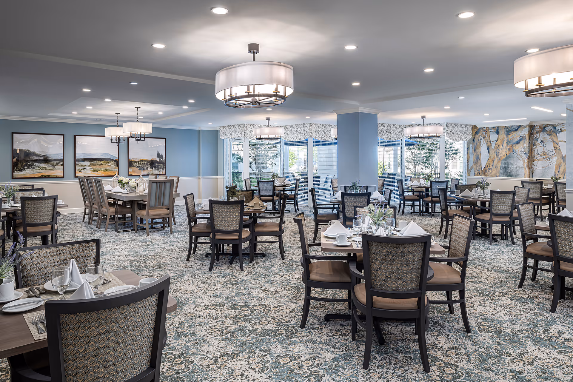 A spacious and elegantly decorated dining room with multiple tables and chairs arranged neatly. The tables are set with white napkins, glassware, and plates. The room features soft blue walls, large windows with blinds, patterned carpet, and modern light fixtures hanging from the ceiling. There are framed landscape paintings on one wall and a large mural of trees on another.