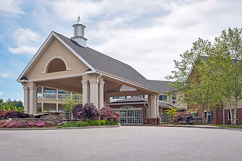 Front exterior view of Lake Pointe Landing Retirement Village featuring a large covered entrance with columns, landscaped flower beds, and trees under a partly cloudy sky.