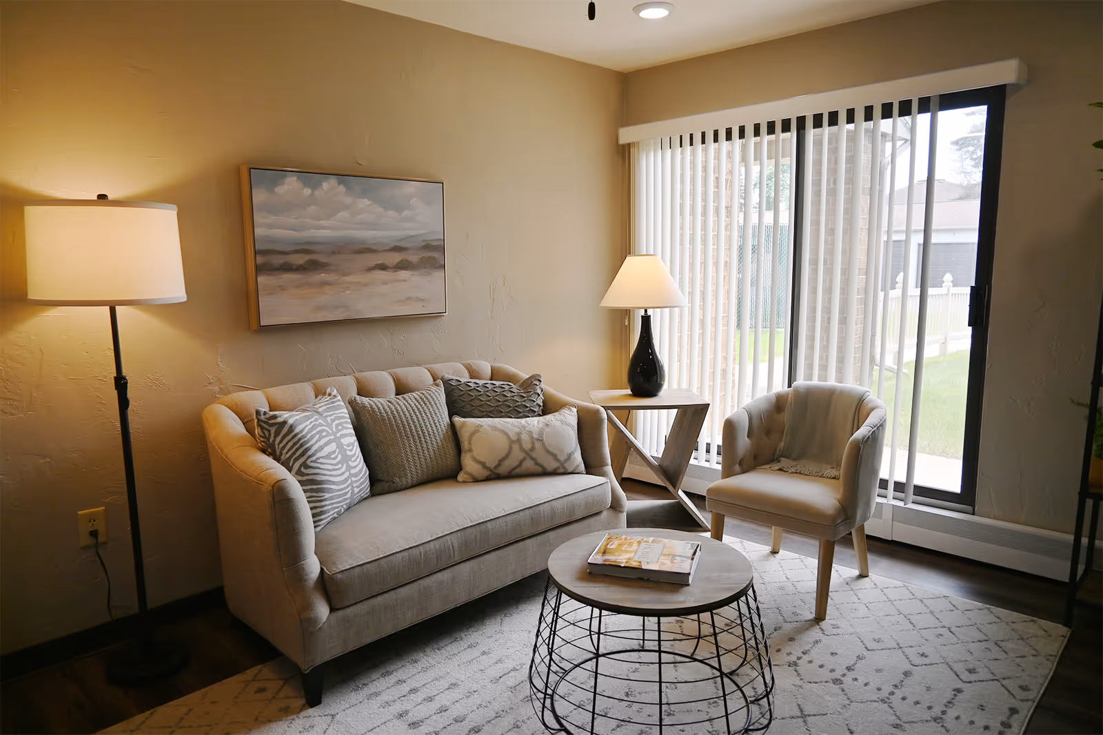 A cozy living room with a beige tufted sofa adorned with patterned pillows, a round coffee table with a book on it, a beige armchair, a floor lamp, a table lamp on a wooden side table, and vertical blinds covering a sliding glass door that leads outside.