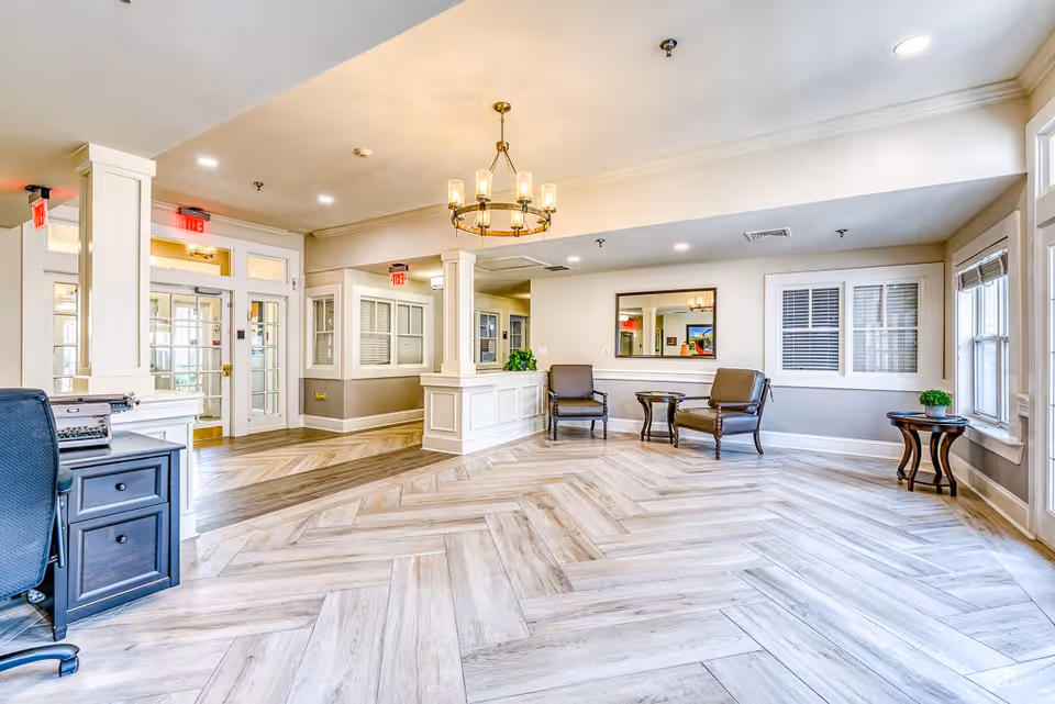 Bright and spacious senior living facility lobby with light wood herringbone flooring, white walls, and large windows letting in natural light. The room features a chandelier, two armchairs with a small round table between them, a reception desk, and a black office chair with a desk on the left side.