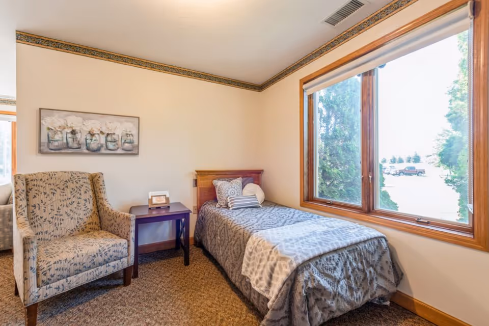A cozy bedroom in a senior living facility featuring a single bed with patterned bedding and multiple pillows, a cushioned armchair with a floral design, a small wooden side table with a framed photo, a large window with a view of trees and parked cars outside, and a floral painting on the wall.
