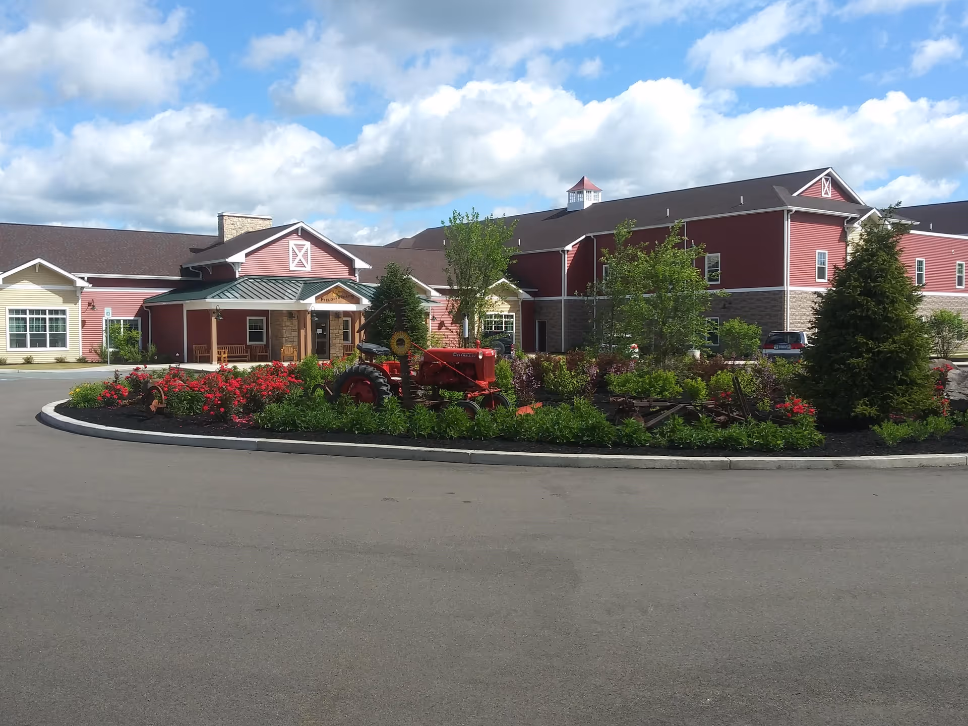 Exterior view of the Field of Dreams facility showing a large red and beige building with a green metal roof entrance. In front of the building is a landscaped roundabout with red flowers, green shrubs, trees, and a vintage red tractor. The sky is partly cloudy.