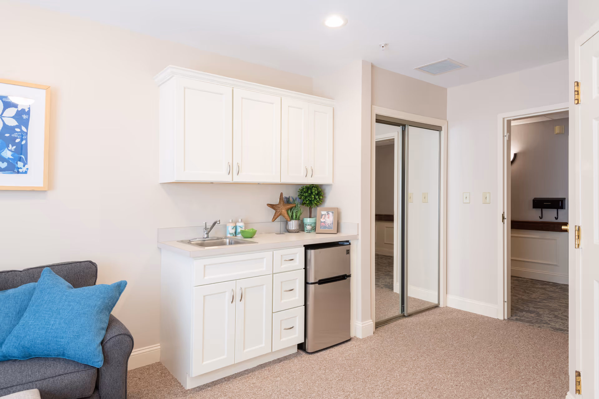 A small kitchenette area with white cabinets, a stainless steel mini refrigerator, a sink, and decorative items including a starfish, a small plant, and a framed photo. To the left is a gray couch with blue pillows, and to the right is a doorway leading to another room with mirrored closet doors.