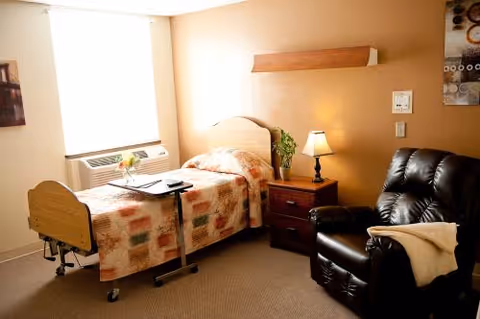 A cozy room with a single hospital-style bed covered with a patterned quilt, a small bedside table with a lamp and a plant, a black recliner chair with a blanket draped over the arm, and a window with a white blind letting in natural light.