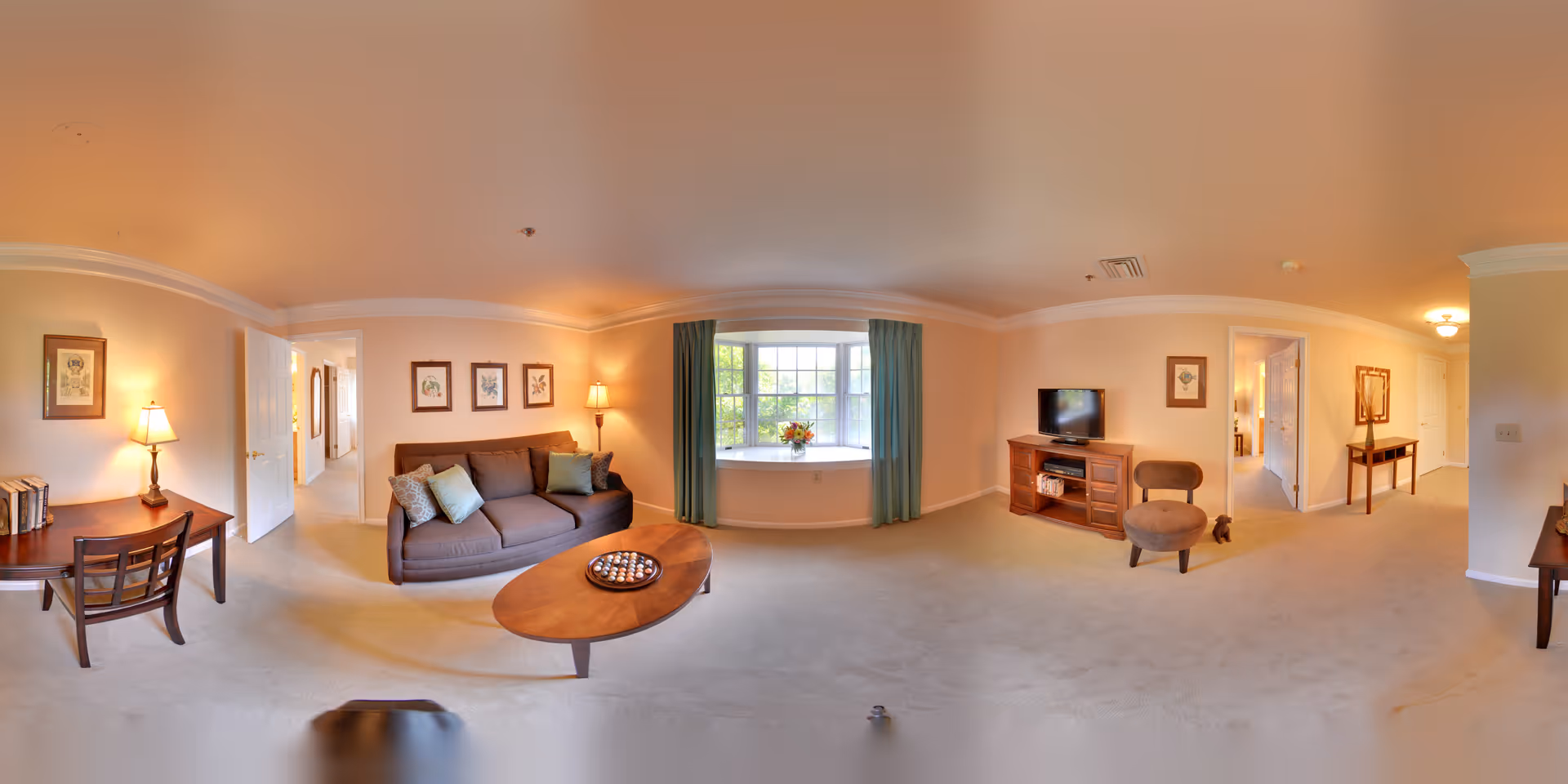 A spacious living room in a senior living facility with beige walls and carpet. The room features a brown sofa with cushions, a wooden coffee table with a decorative tray, a wooden TV stand with a flat-screen TV, a small upholstered chair, and a desk with a chair and lamp. There is a large window with green curtains and a flower arrangement on the windowsill. The room is softly lit with table lamps and ceiling lights, and doorways lead to other rooms.