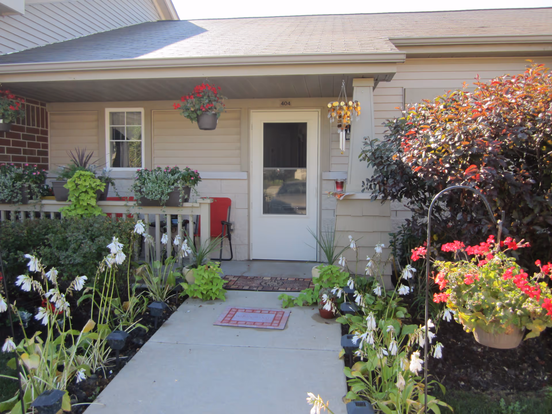 Front entrance of a senior living facility unit with a white door numbered 404, surrounded by a small porch with hanging flower pots, a red chair, and various plants and flowers along the walkway leading to the door.
