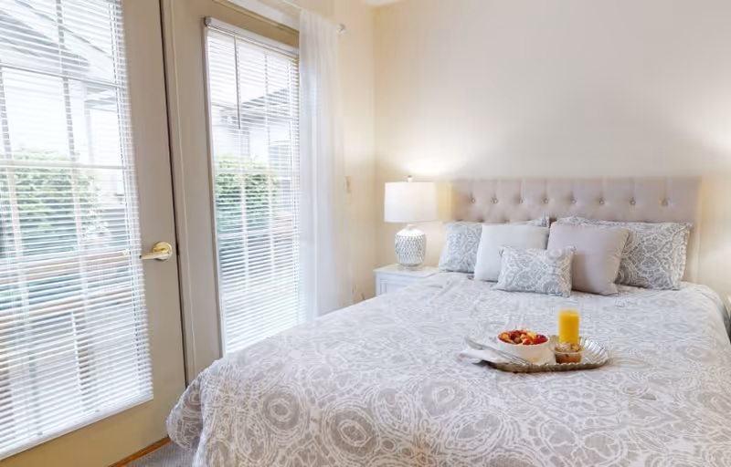 A bright bedroom with a large bed covered in a patterned white and gray bedspread, several pillows, a bedside table with a decorative lamp, and a tray with a bowl of fruit and a glass of orange juice on the bed. There are glass doors with blinds and sheer white curtains letting in natural light.