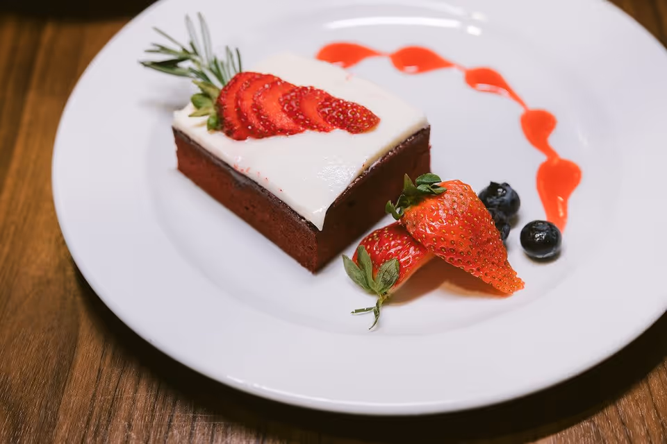 A square piece of red velvet cake topped with white frosting and sliced strawberries, garnished with a sprig of rosemary. The plate also has whole strawberries, blueberries, and a decorative swirl of red sauce.