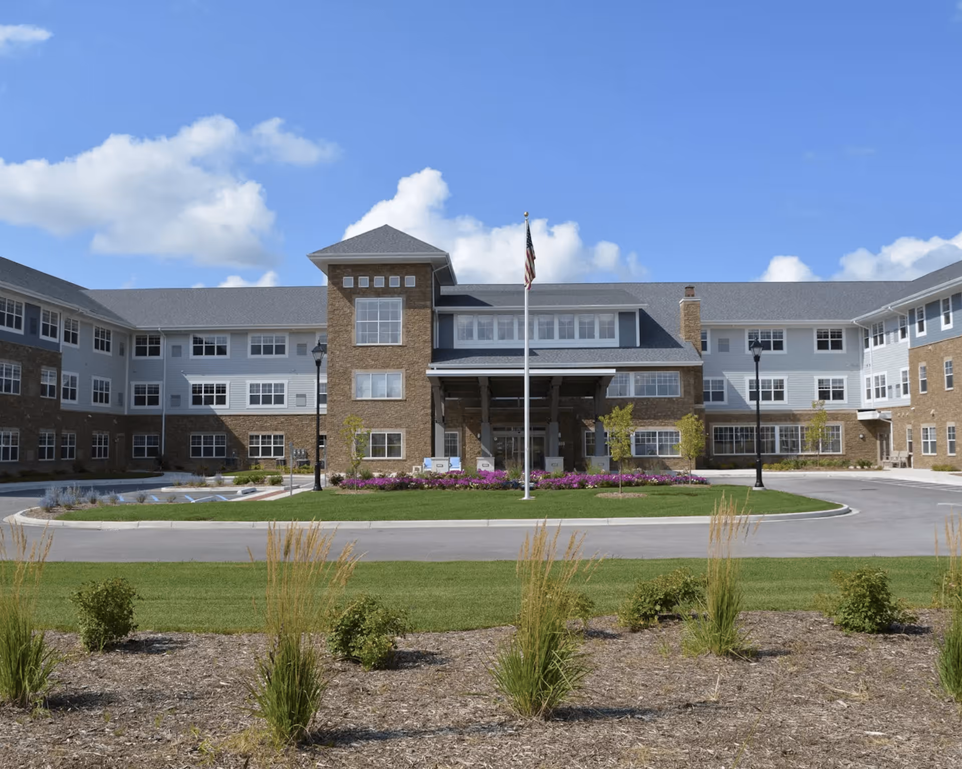 Front exterior view of a large senior living facility building with a circular driveway, landscaped garden with grass, bushes, and flowers, and an American flag on a flagpole in front. The building has multiple windows, a covered entrance, and a combination of brick and siding exterior under a blue sky with some clouds.