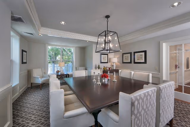 Bright formal dining room with a large dark wood table surrounded by upholstered chairs, a central chandelier, framed artwork and a window showing greenery.
