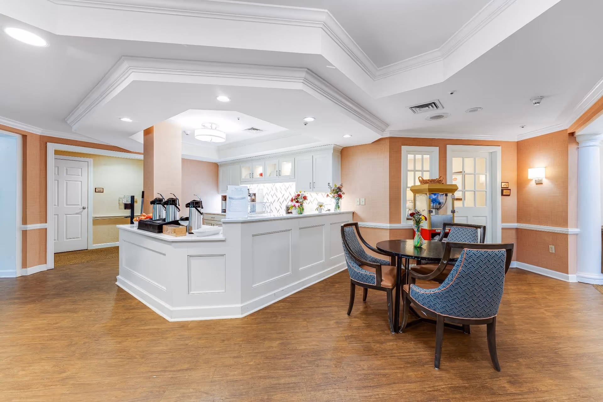 A bright and clean interior space featuring a white service counter with coffee dispensers and a small table with four upholstered chairs. The room has warm peach-colored walls, wood flooring, and white trim. There is a popcorn machine in the background near double glass doors, and ceiling lights provide ample illumination.