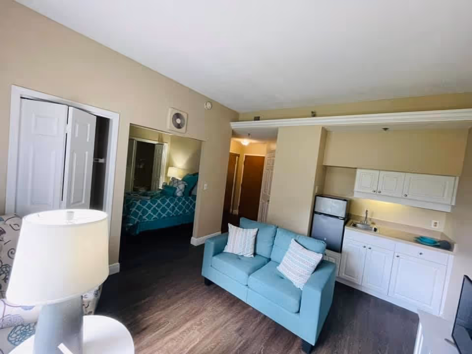 Interior view of a senior living facility unit showing a small living area with a light blue loveseat with two pillows, a kitchenette with white cabinets, a small refrigerator, and a sink. To the left, there is a glimpse of a bedroom with a bed covered in a blue and white patterned bedspread. A lamp and an armchair are partially visible in the foreground.