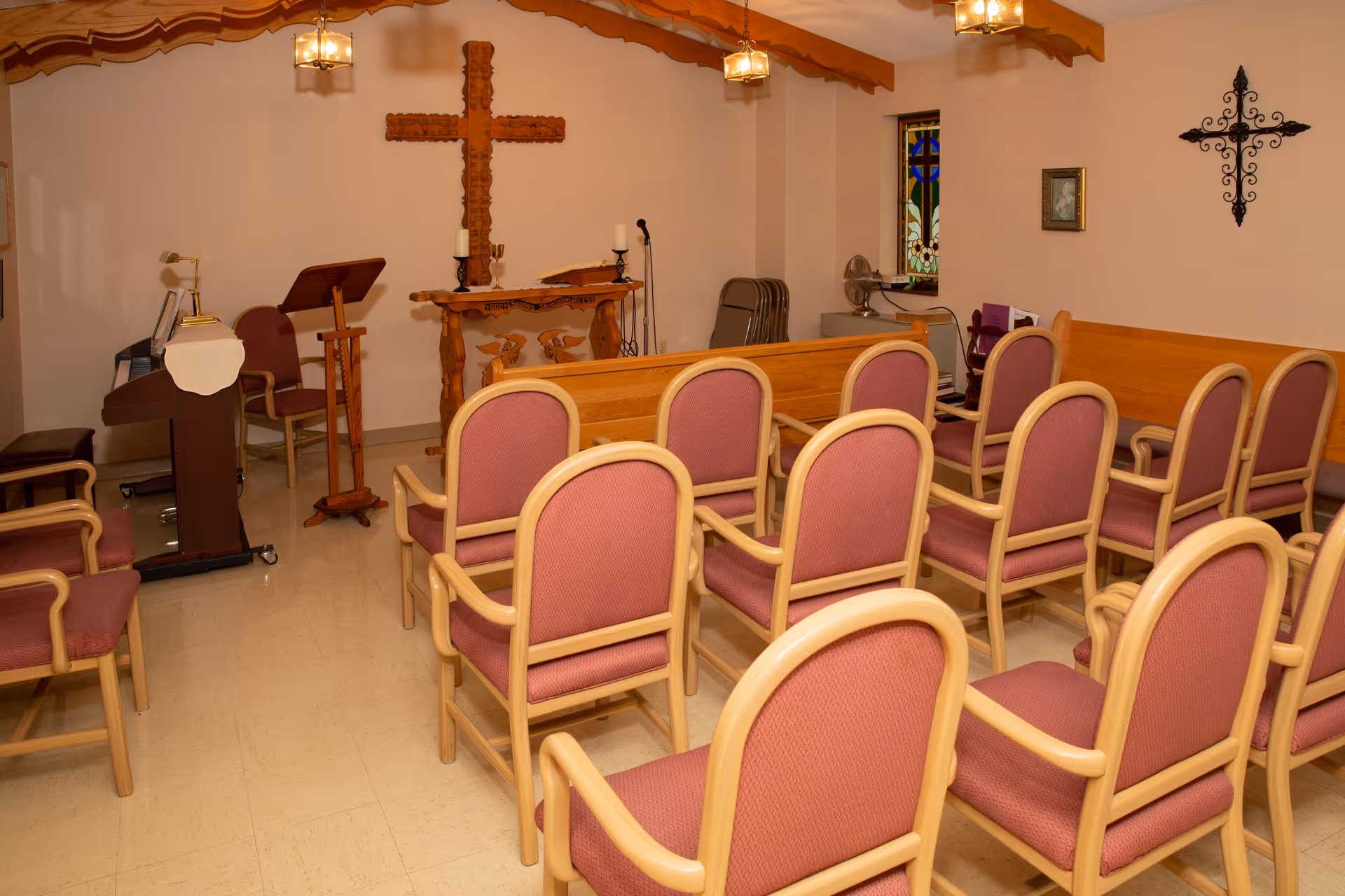 Interior of a small chapel or prayer room with rows of wooden chairs with pink cushions facing a wooden altar with a large wooden cross on the wall behind it. There are two candle holders on the altar, a lectern, a microphone stand, a stained glass window, and decorative wall crosses.