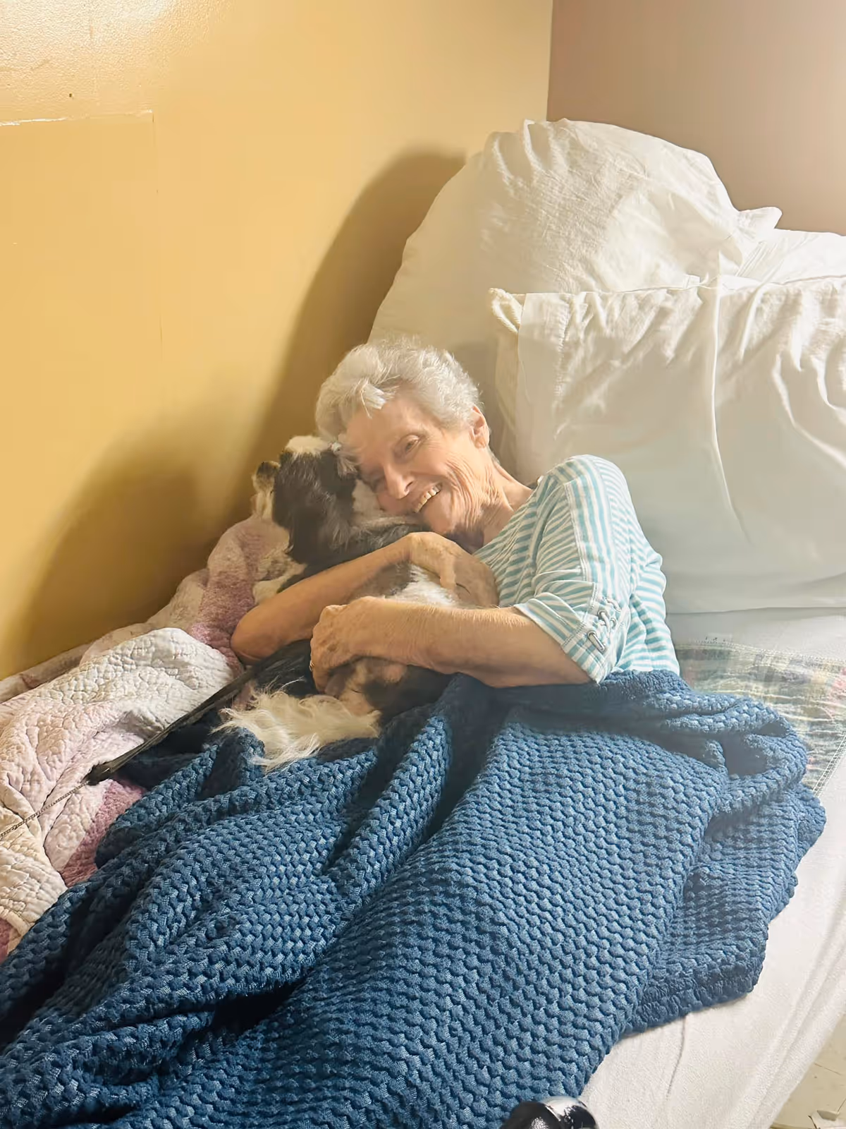 An elderly woman lying in bed, smiling and hugging a small dog. The bed has white pillows and a blue knitted blanket. The background wall is painted yellow.