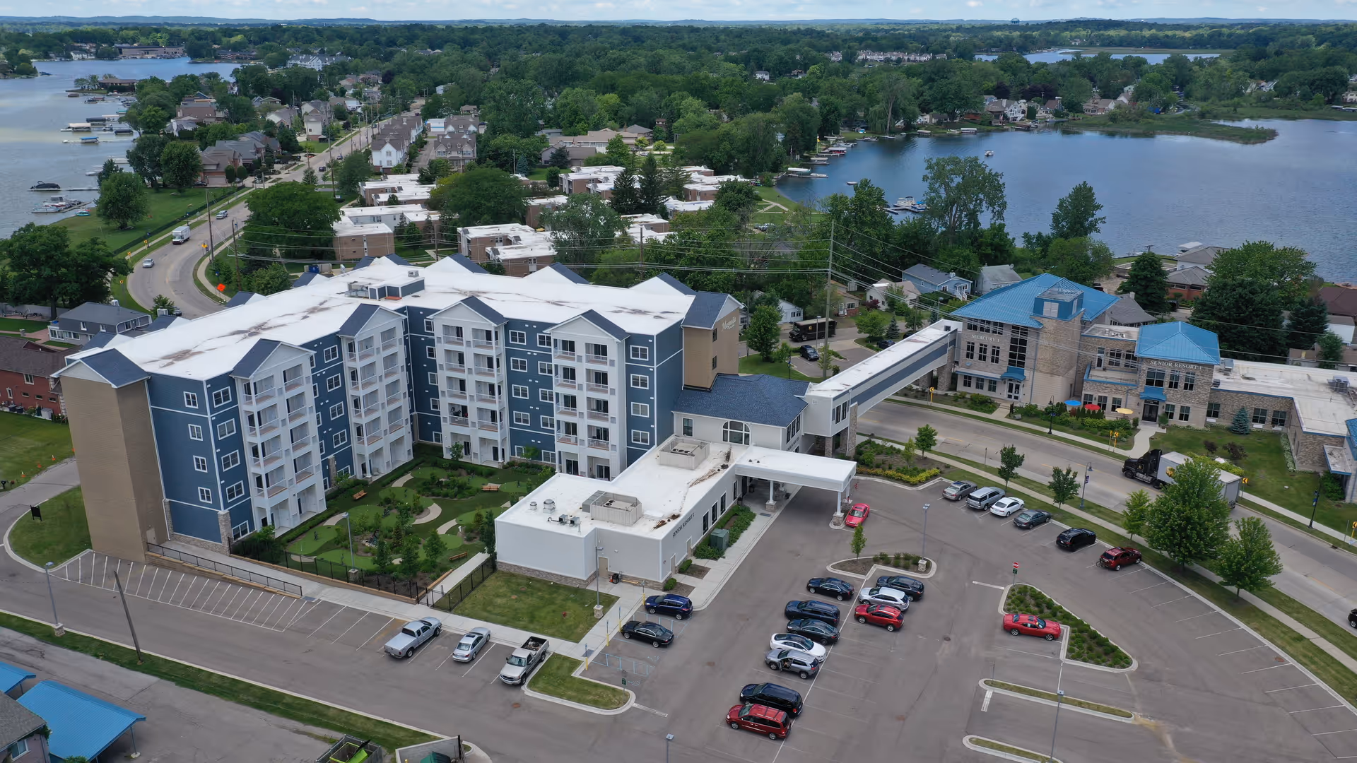 Aerial view of a multi-story senior living building with a parking lot, skybridge, and nearby lake.