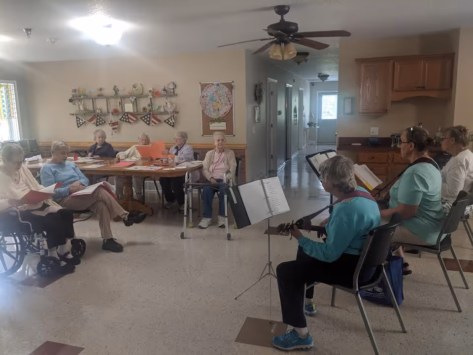 A group of elderly women seated in a common room at Woodland Acres Assisted Living. Some women are sitting around a table with papers, while others are seated in chairs facing two women playing guitars and singing from music stands. The room has beige walls, wooden cabinets, and a ceiling fan.