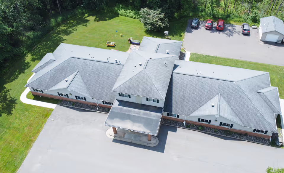 Aerial view of a single-story senior living facility building with a covered entrance, surrounded by green lawn and trees. There is a parking area with several cars and a small detached garage or shed nearby.