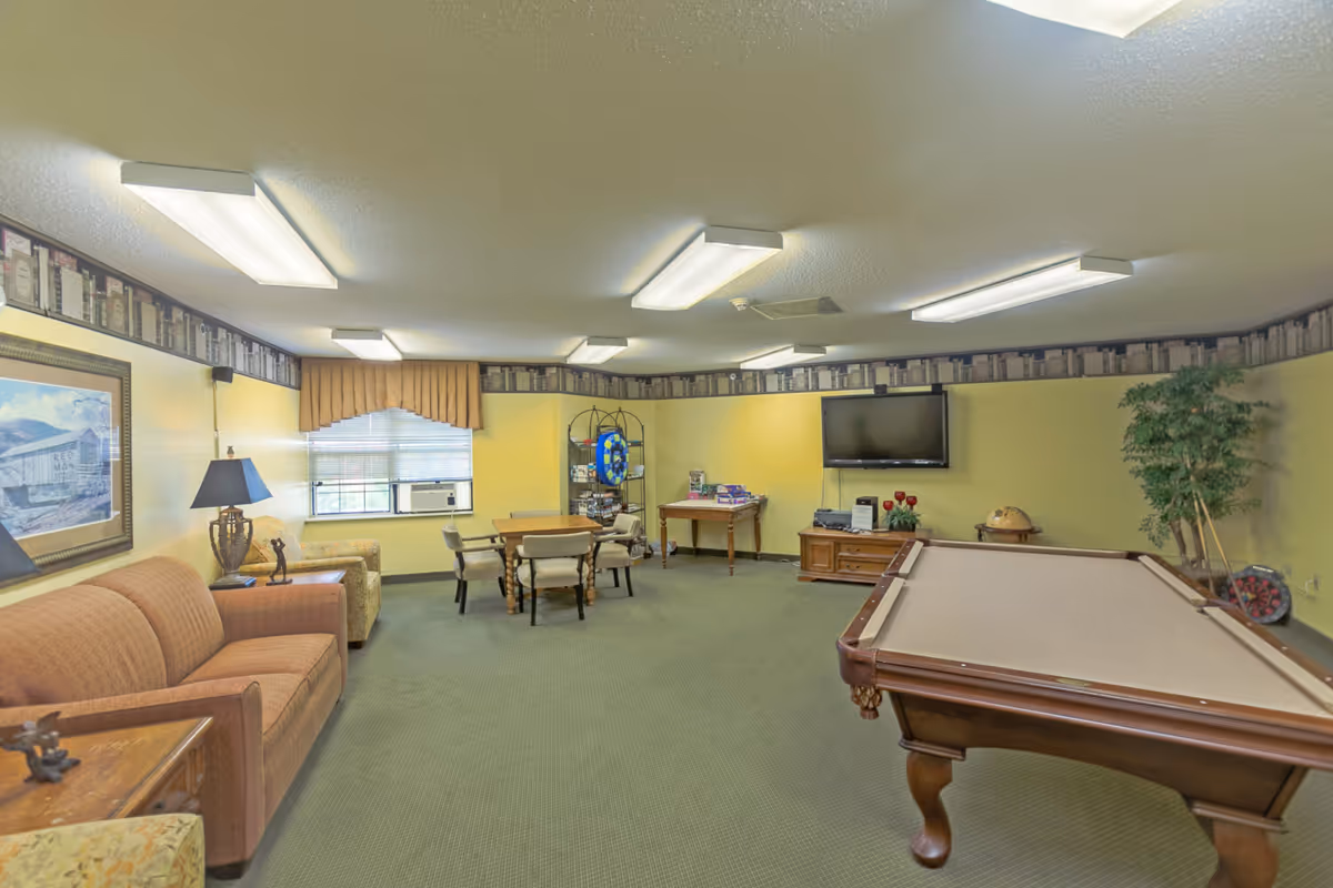 A recreational room with a pool table, a flat-screen TV mounted on a yellow wall, a wooden cabinet, a table with board games, a round table with chairs, a sofa, an armchair, a window with blinds and a valance, and a potted plant in the corner. The room has fluorescent ceiling lights and a carpeted floor.