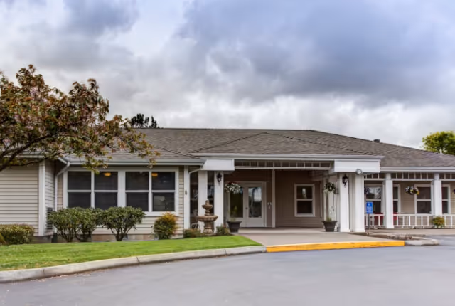 Exterior view of a single-story senior living facility building with a covered entrance, beige siding, and a gray shingled roof. There are shrubs and small trees in front of the building, a paved driveway, and a cloudy sky overhead.