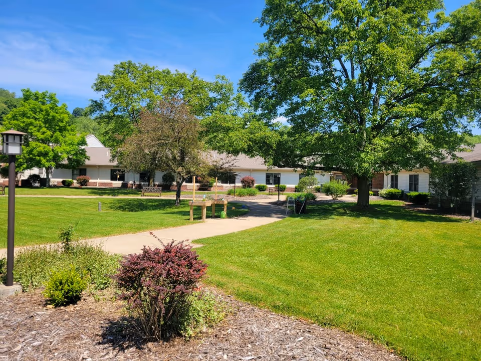 A sunny outdoor garden area at Sugar Creek Care Center featuring a well-maintained lawn, several trees providing shade, a paved walkway, and some bushes and plants in the foreground. The building is visible in the background with white siding and multiple windows.