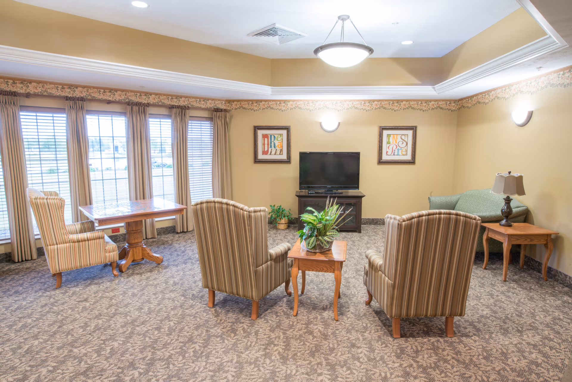 A cozy living room area in a senior living facility with three striped armchairs arranged around a small wooden table with a plant on it. There is a flat-screen TV on a stand against the wall, flanked by two framed artworks. A green loveseat and a wooden side table with a lamp are positioned in the corner. Large windows with beige curtains allow natural light to fill the room, which has beige walls and patterned carpet.