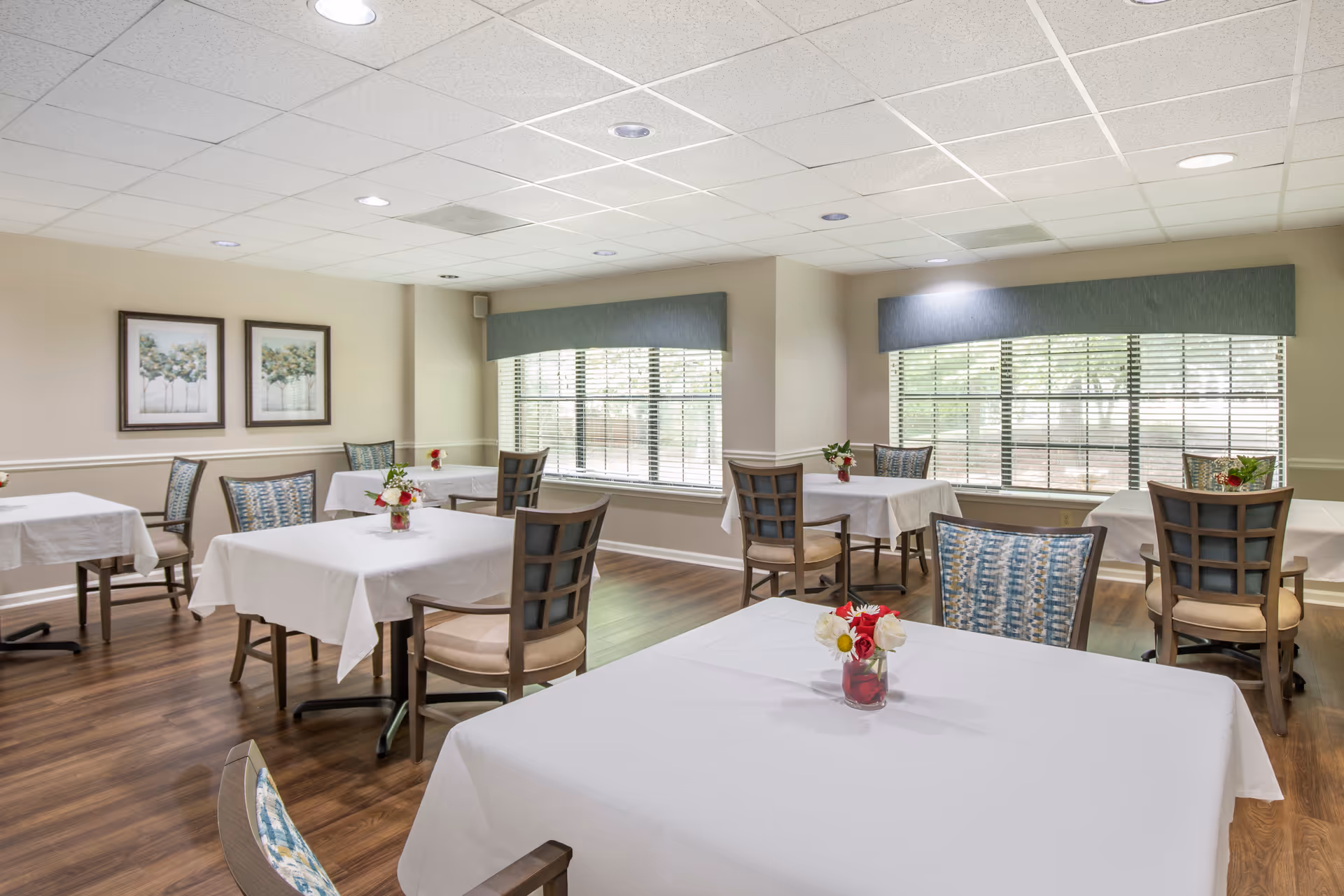 A bright dining room with several tables covered in white tablecloths, each adorned with a small vase of flowers. The room has wooden floors, large windows with blinds and valances, and framed artwork on the walls. Chairs with patterned upholstery surround the tables.