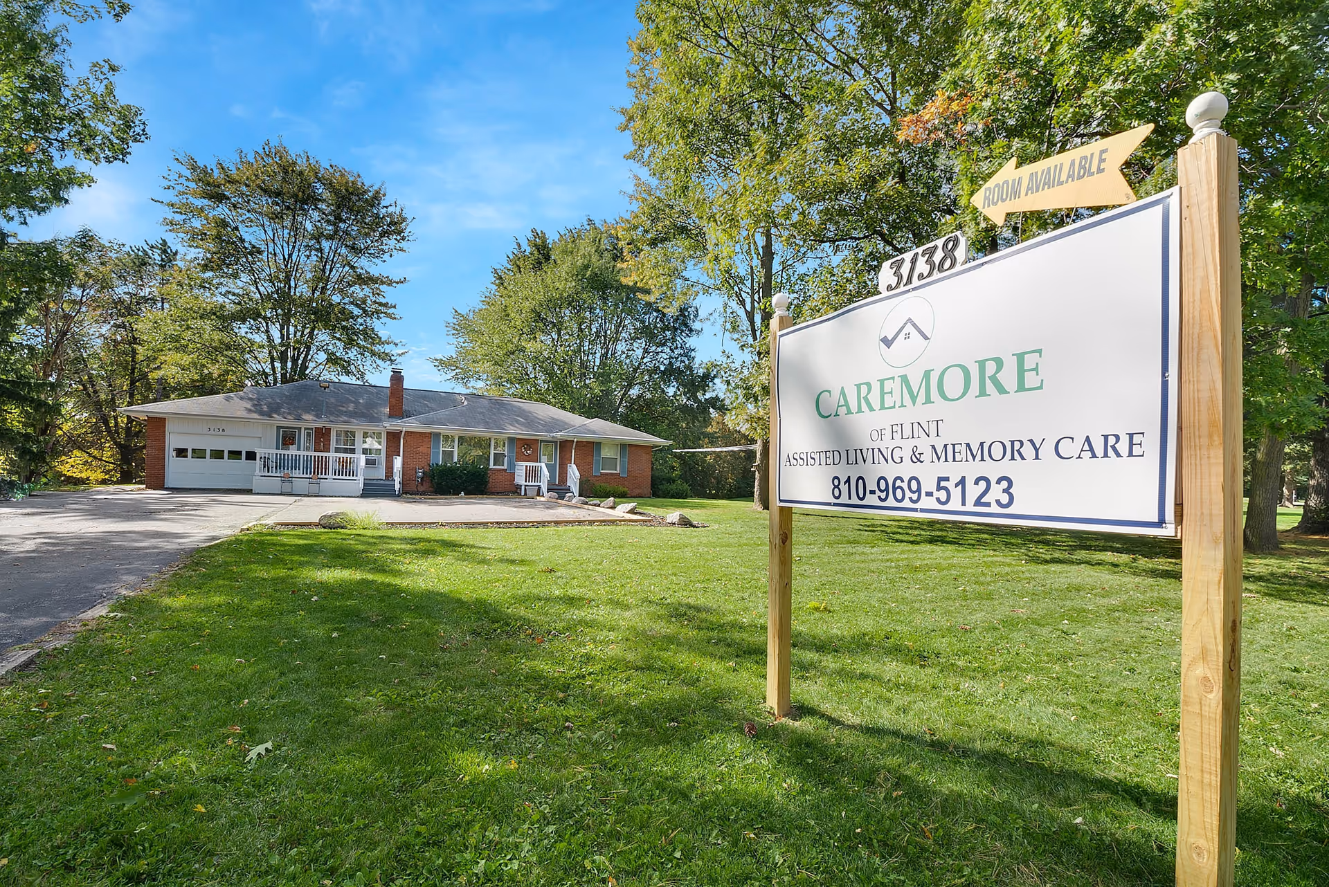 Exterior view of a single-story brick building with a driveway and a green lawn. A large sign in the foreground reads 'Caremore of Flint Assisted Living & Memory Care' with a phone number and an arrow sign indicating 'Room Available'. Trees with green foliage are visible in the background under a blue sky.