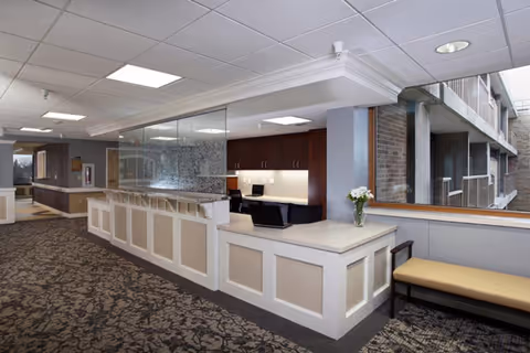 Lobby reception area with a long paneled front desk, office workspace behind glass, patterned carpet, and bench seating by a window.