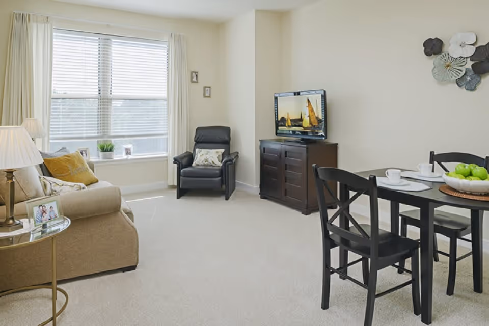 A bright living room with a beige sofa adorned with cushions, a black armchair, a dark wooden TV stand with a flat-screen TV displaying a sailboat scene, and a black dining table set with four chairs, white cups, and a bowl of green apples. The room has a large window with white blinds and curtains, light-colored walls, and carpeted flooring.